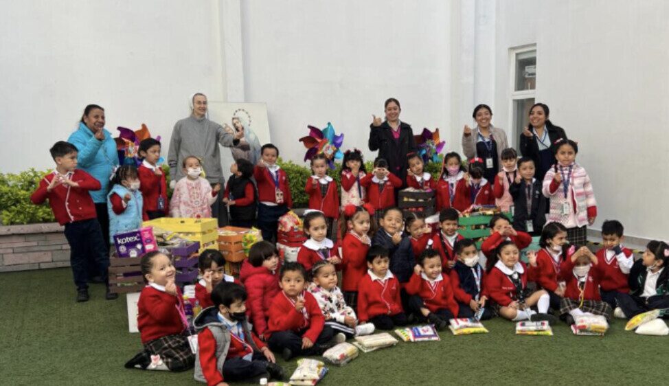 Un grupo numeroso de niños de preescolar posa en el patio del colegio, junto a una hermana y varias maestras, rodeados de cajas y paquetes reunidos en una colecta.
