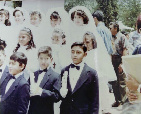 Niños de primera comunión, de traje y con velas en la mano, ante un mural con los retratos de sus compañeras.
