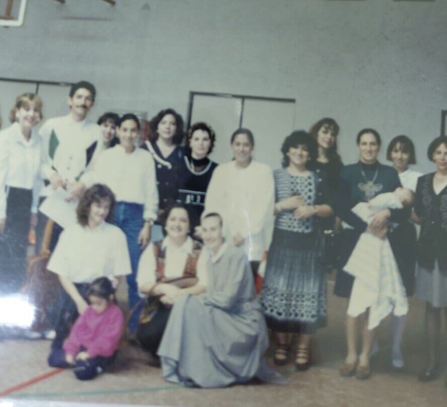 Foto de grupo en un salón: una hermana marcelina, de hábito gris y al frente, rodeada de mujeres y niños.