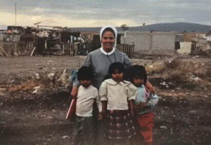 Sor María Rosa de pie con tres niños en un descampado de la colonia Bolaños, con casas de lámina al fondo.